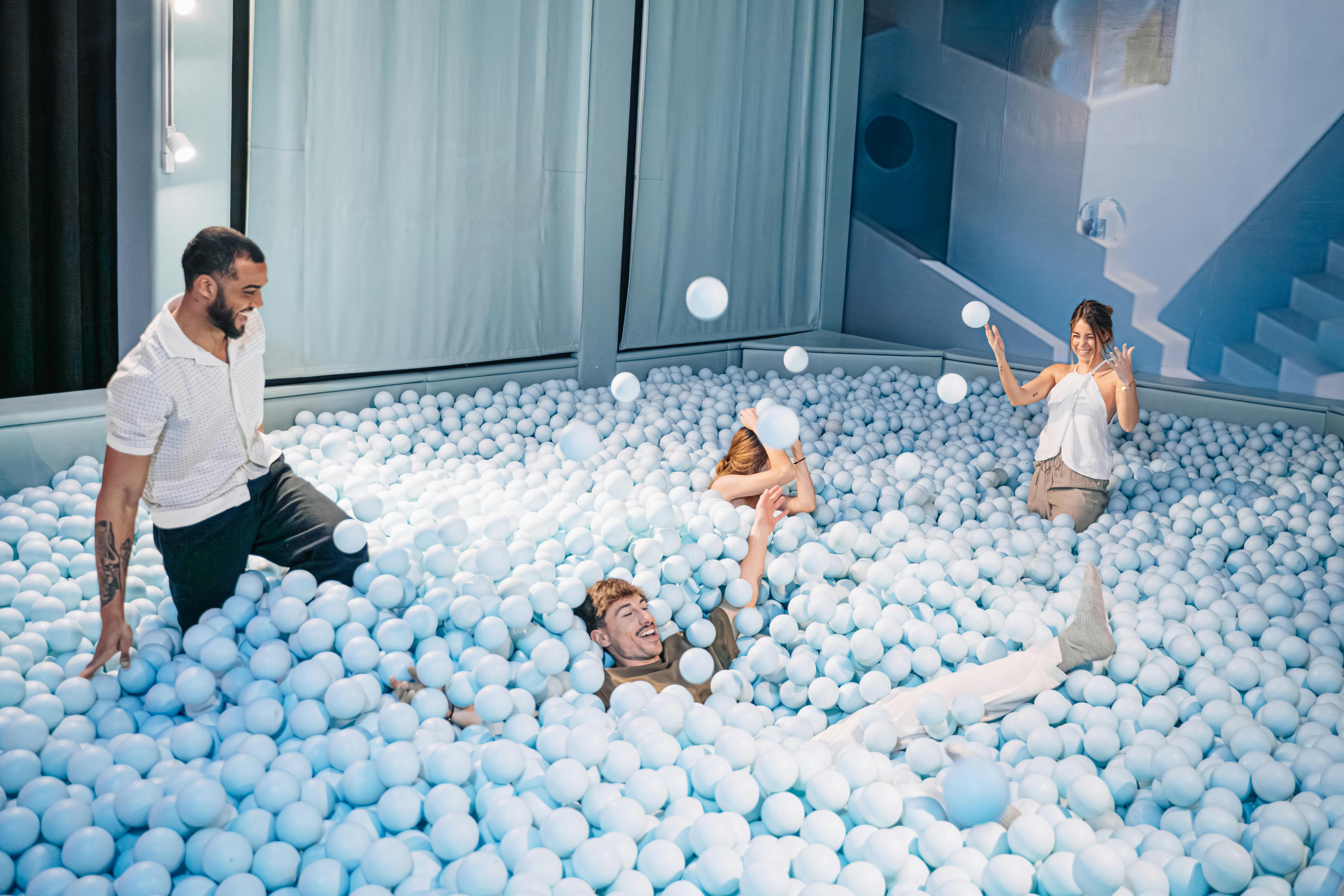 四个成年人在一个充满浅蓝色塑料球的大型游泳池里玩耍，周围是浅蓝色的墙壁和窗帘。