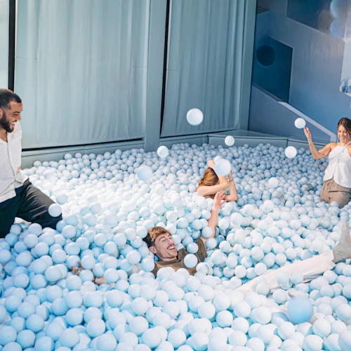 Fyra vuxna leker glatt i en stor pool fylld med ljusblå plastkulor, omgiven av ljusblå väggar och gardiner.