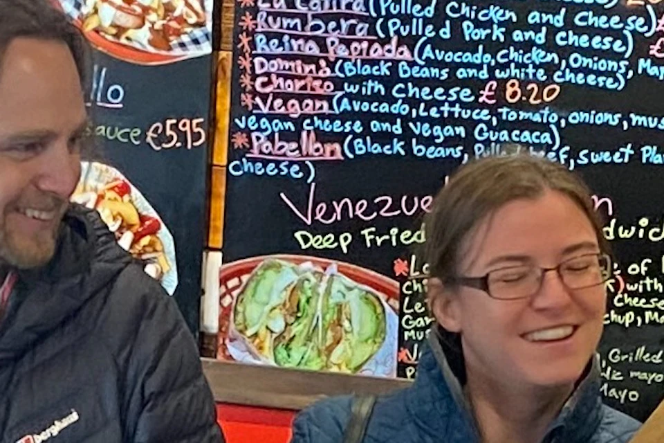 Four people stand at a counter, smiling and chatting with an employee in a cozy cafe featuring a wooden backdrop and chalkboard menu.