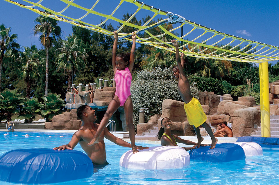 Two children hang from a rope bridge over a pool, with an adult and another child on inflatables beneath them.