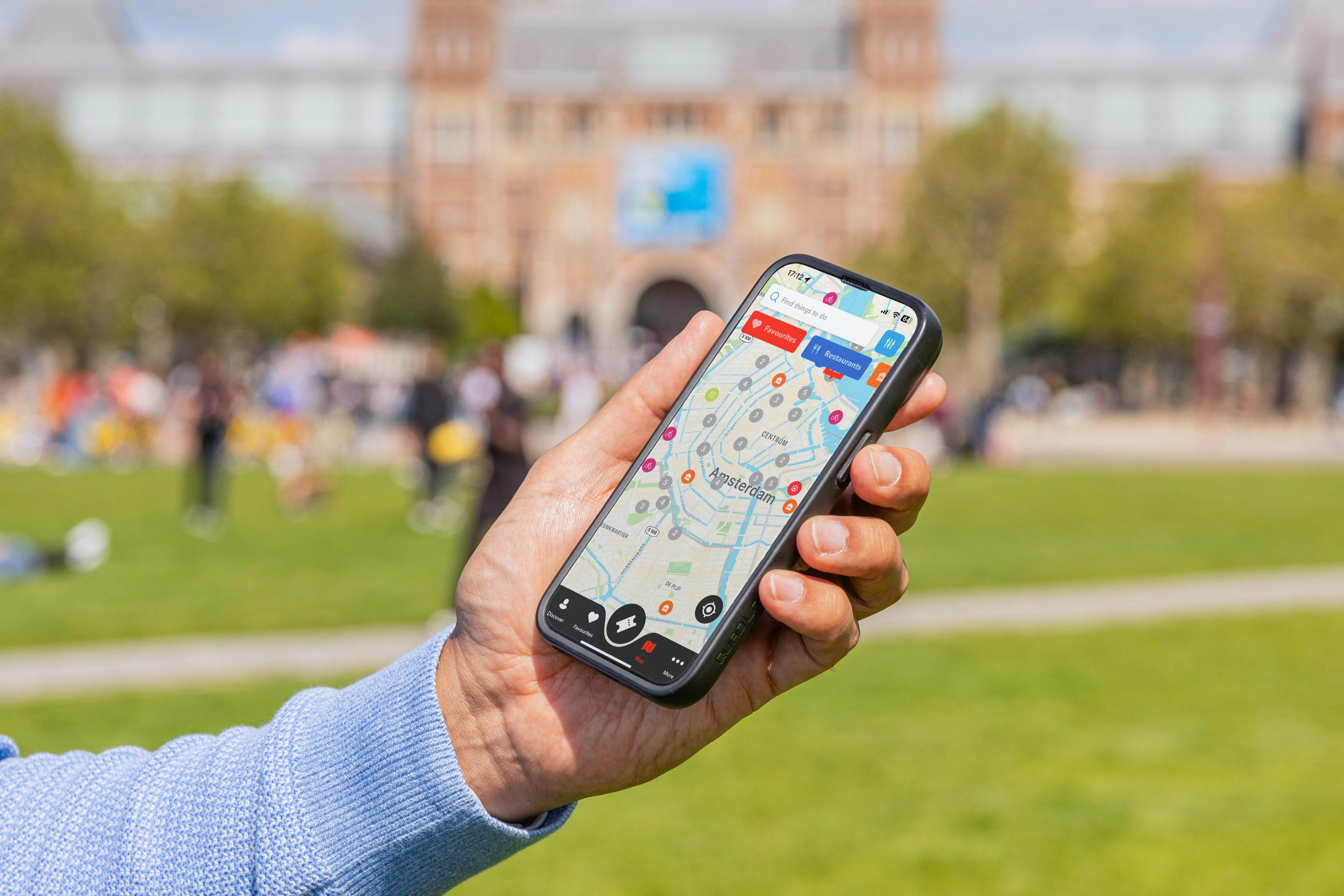 En hand som håller en smartphone som visar en kartapp, med en suddig utomhuspark och byggnad i bakgrunden.