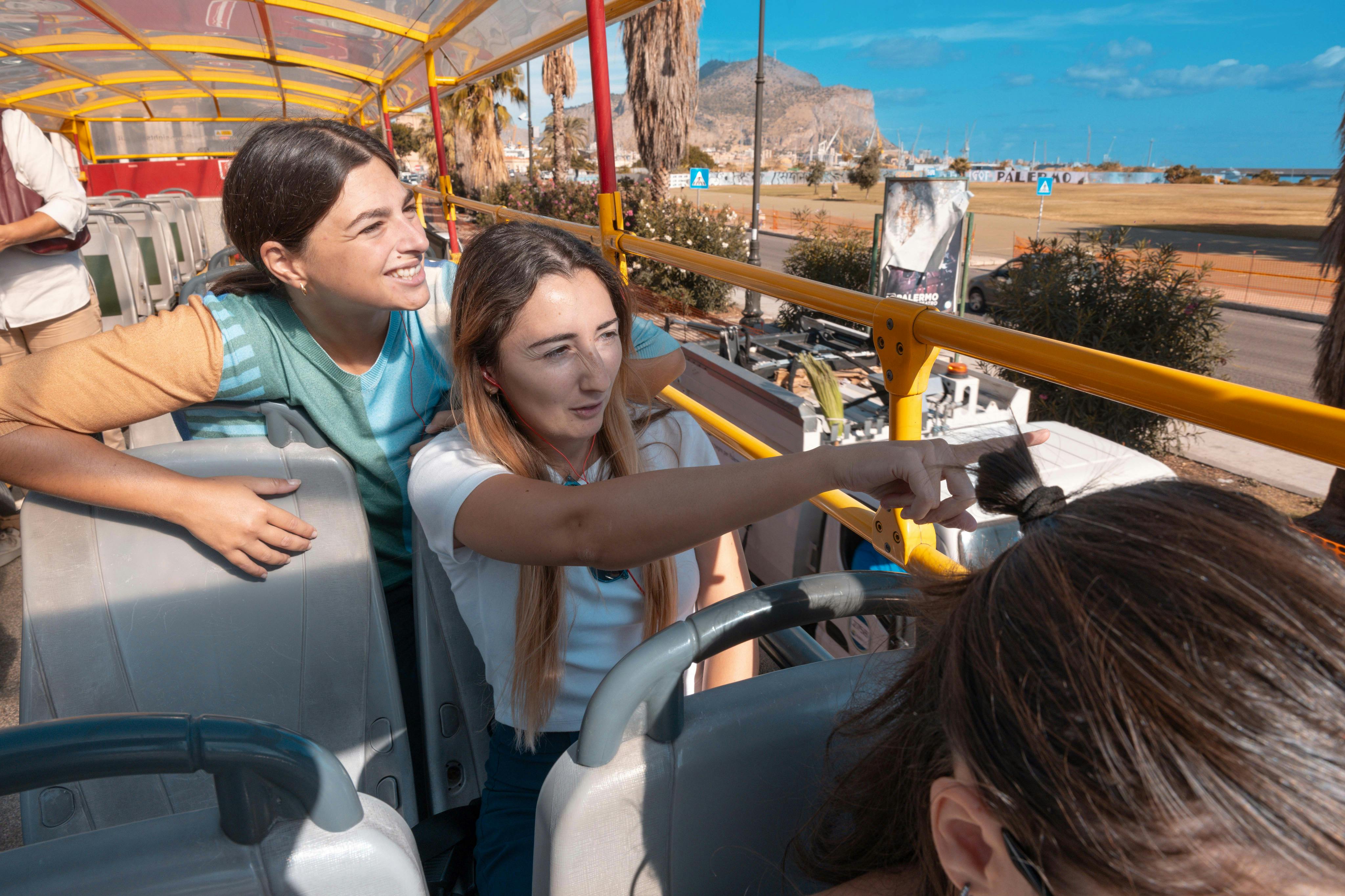 Due donne sedute sul ponte superiore di un autobus scoperto, una indica eccitata mentre l'altra guarda e sorride.