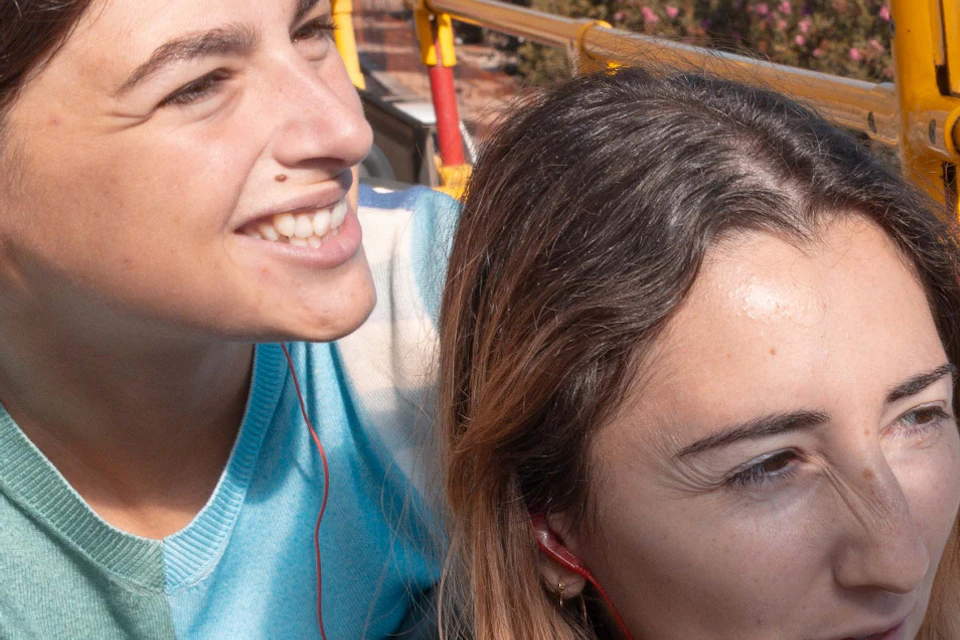 Two women sitting on the upper deck of an open-top bus, one pointing excitedly while the other looks on and smiles.