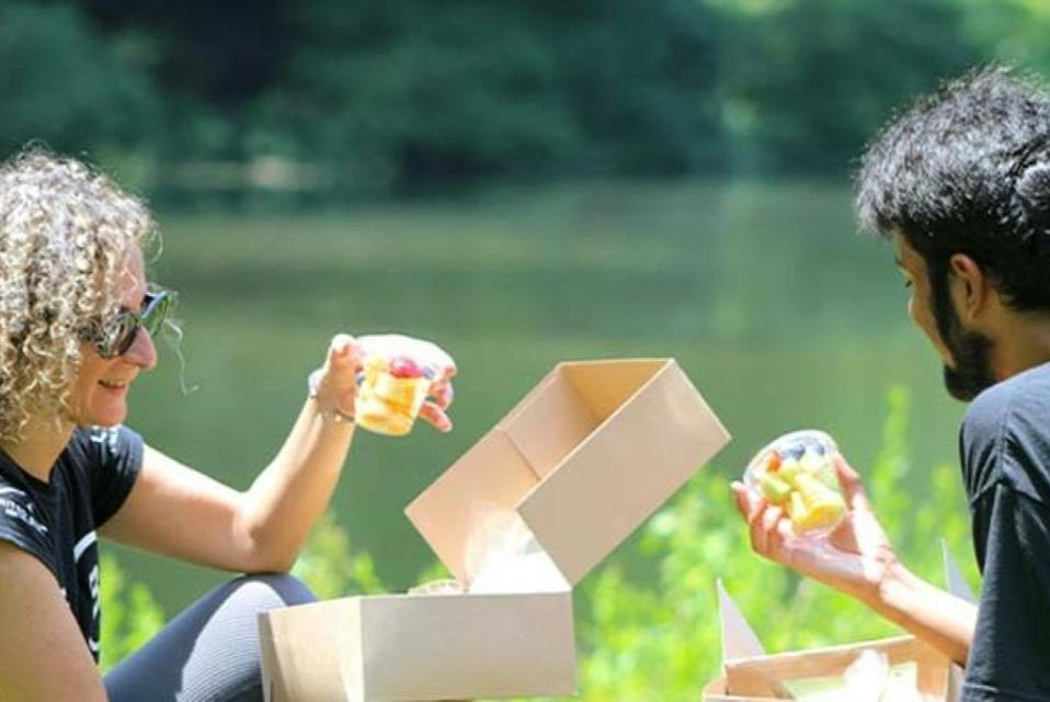 A person with curly hair and sunglasses sits outdoors, holding a drink near an open cardboard box, with greenery and water in the background.