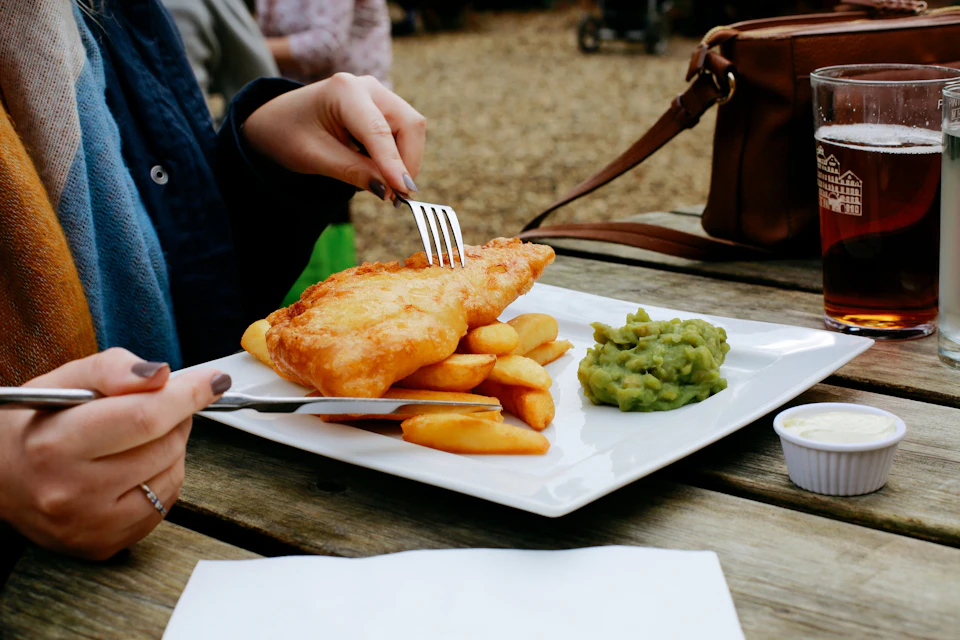 A person eating fish and chips with mushy peas on a white plate outdoors, using a fork and knife, with a bag nearby.