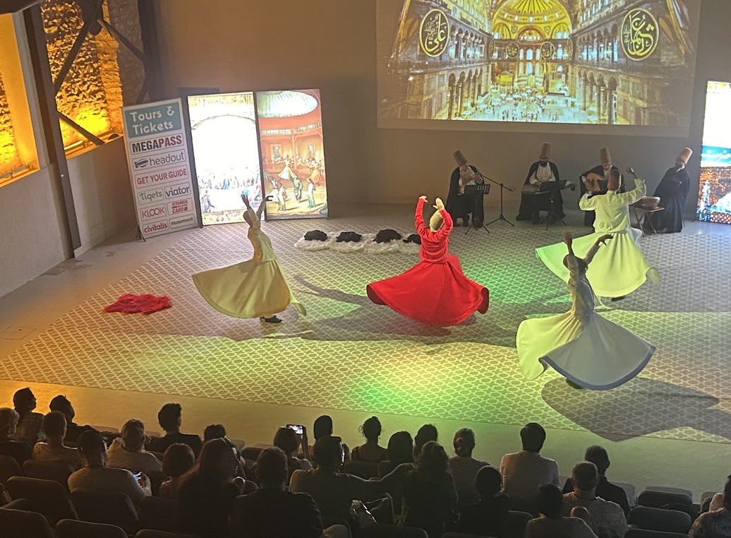 Χορευτές με πολύχρωμες ρόμπες εκτελούν έναν παραδοσιακό περιστρεφόμενο χορό στη σκηνή, με μουσικούς στο παρασκήνιο και το κοινό να παρακολουθεί.