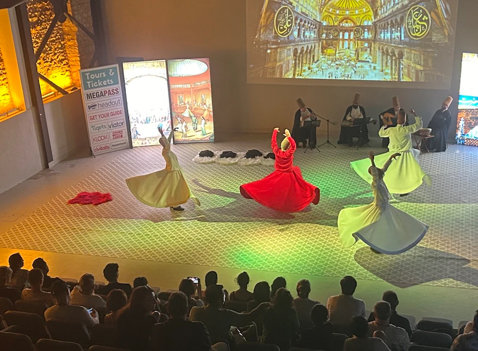 Dancers in colorful robes perform a traditional spinning dance on stage, with musicians in the background and an audience watching.