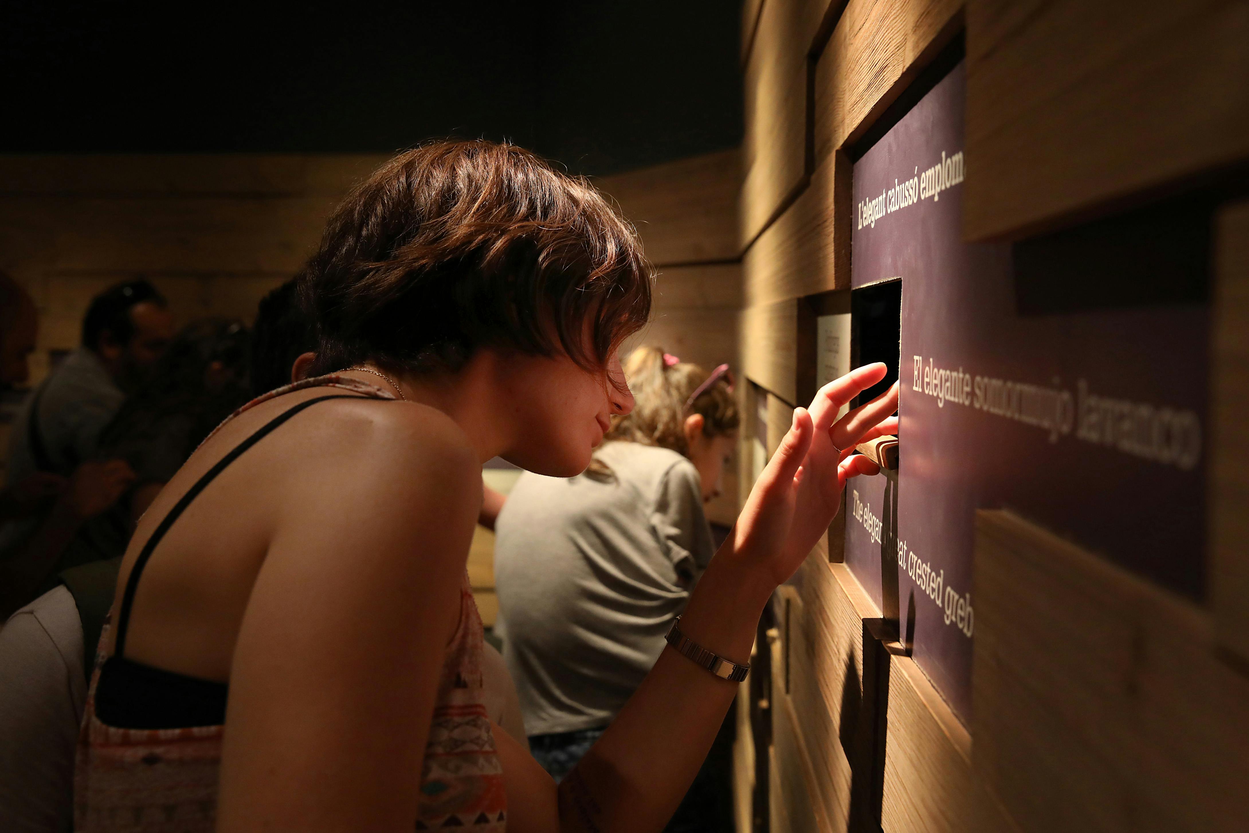 Osoba se dívá malým otvorem v dřevěné stěně s textem, zatímco jiná osoba je v pozadí.