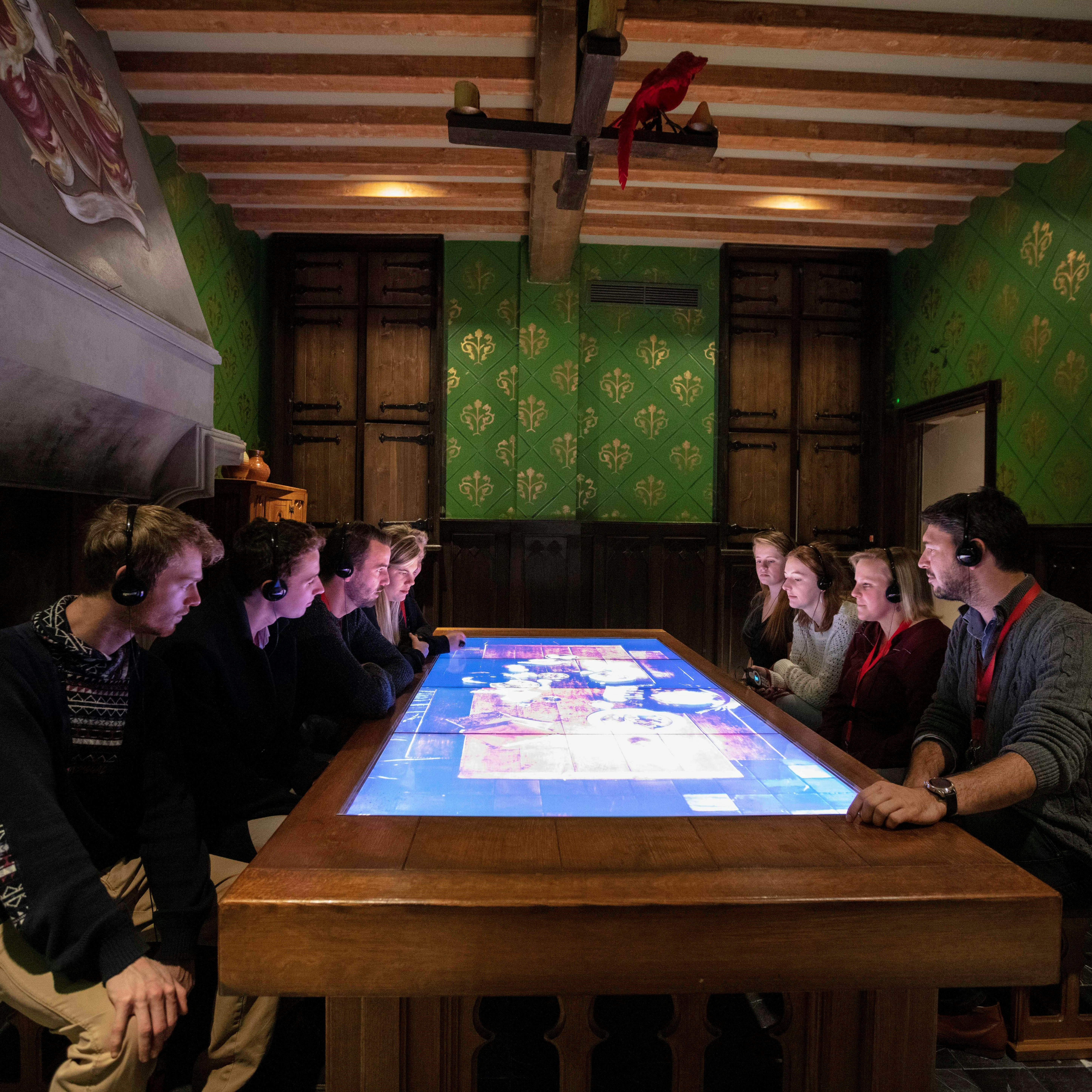 Lidé sedící kolem velkého interaktivního stolu v tlumeně osvětlené místnosti s dřevěnými trámy a zelenými vzorovanými stěnami, se sluchátky na uších.