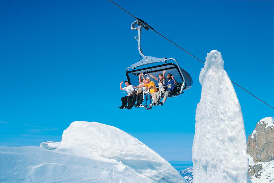 People riding a ski lift above snowy mountains, with clear blue sky and large ice formations in view.