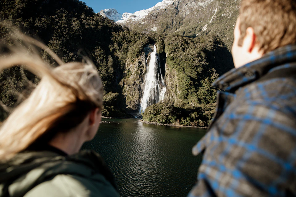 A scenic waterfall flowing down a rocky cliff surrounded by greenery, viewed from behind two people admiring the scene.