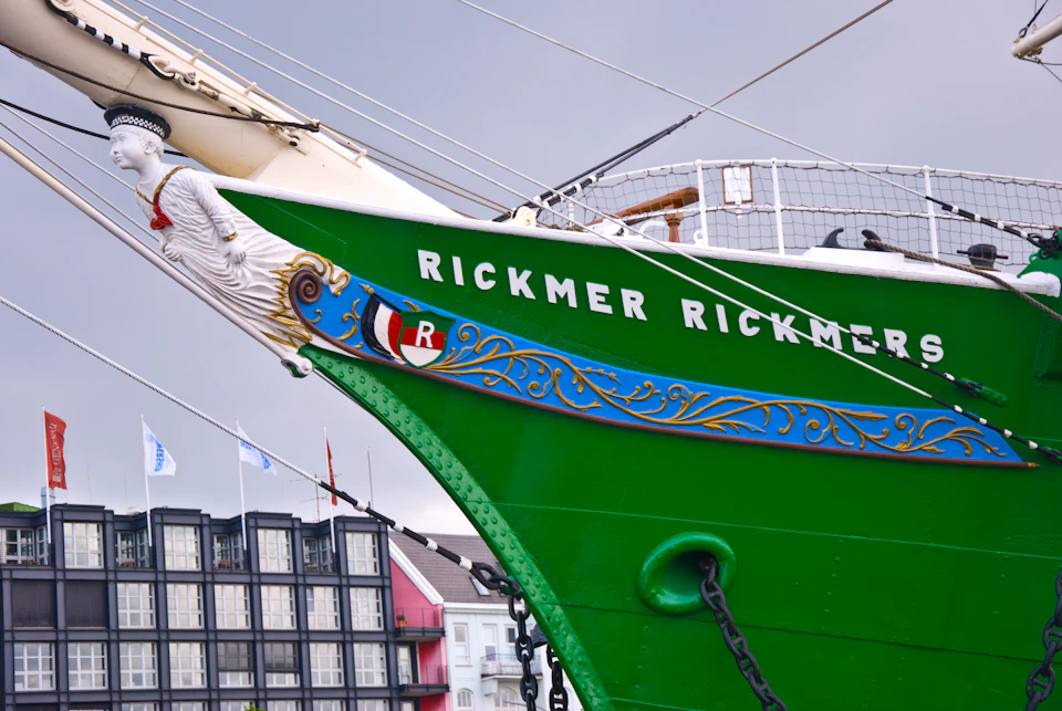 Bow of a green ship named "Rickmer Rickmers" with decorative blue and gold trim, docked near buildings under a gray sky.