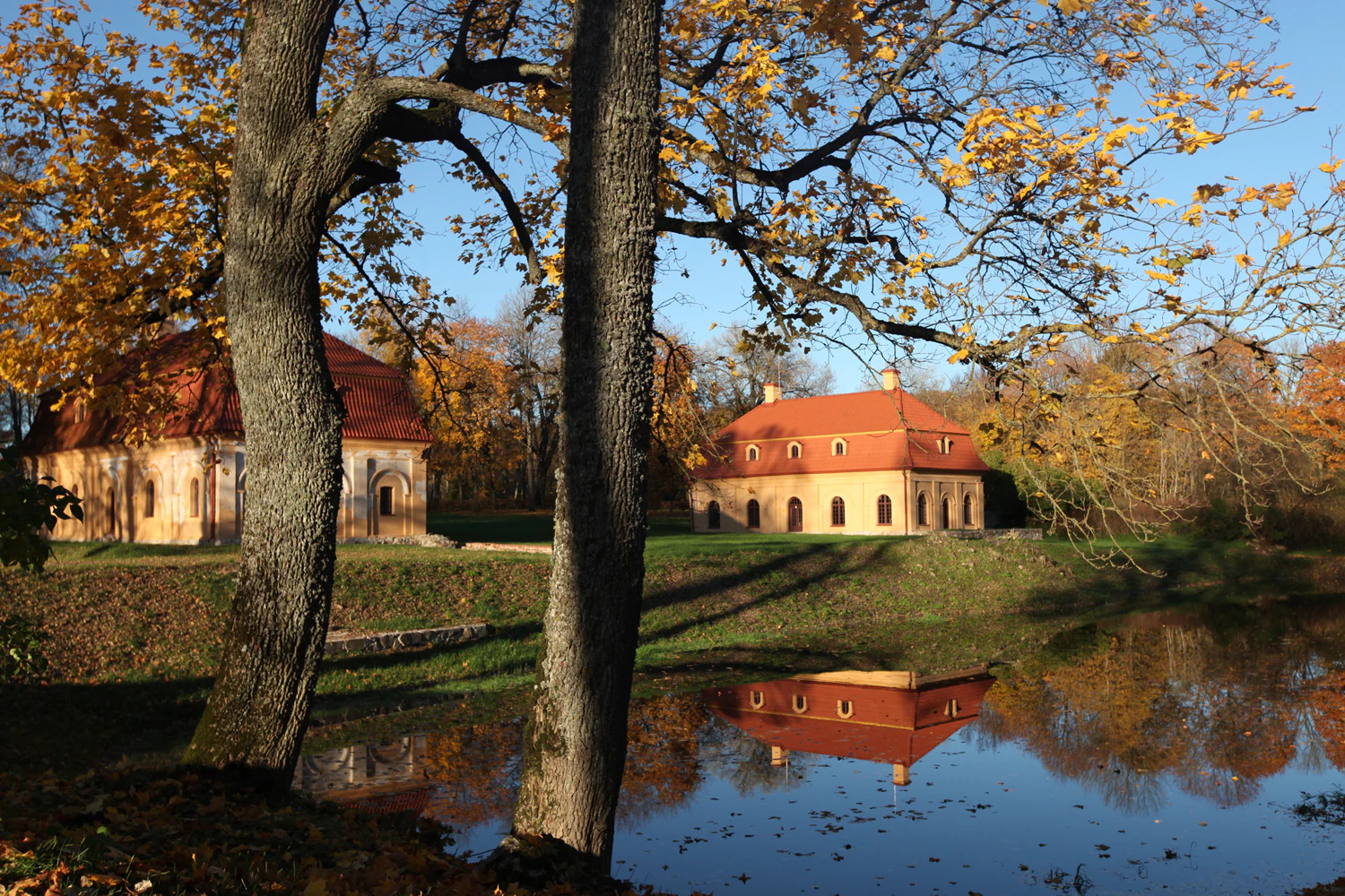 Two trees in the foreground, autumnal building with red roof reflected in a pond, surrounded by fall foliage and a clear sky.