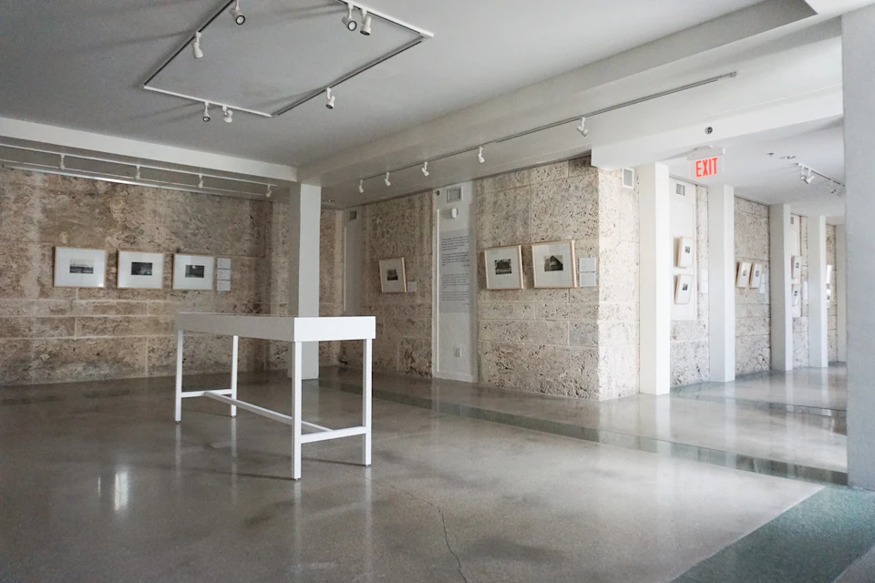 A minimalist art gallery with stone walls, framed artworks, white display tables, and a polished concrete floor.