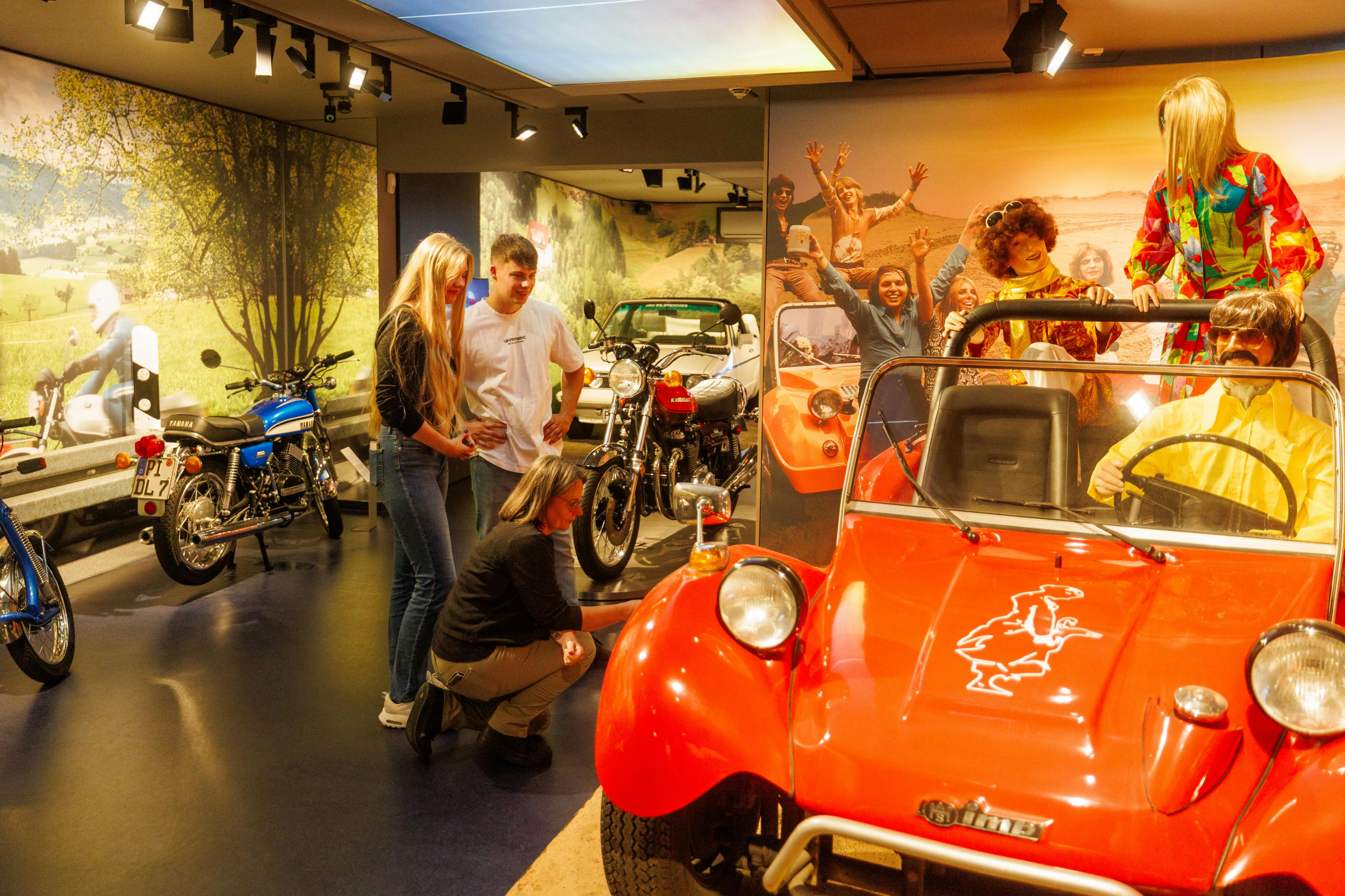 A museum exhibit featuring vintage motorcycles, a red car, and three people observing the displays under bright lighting.