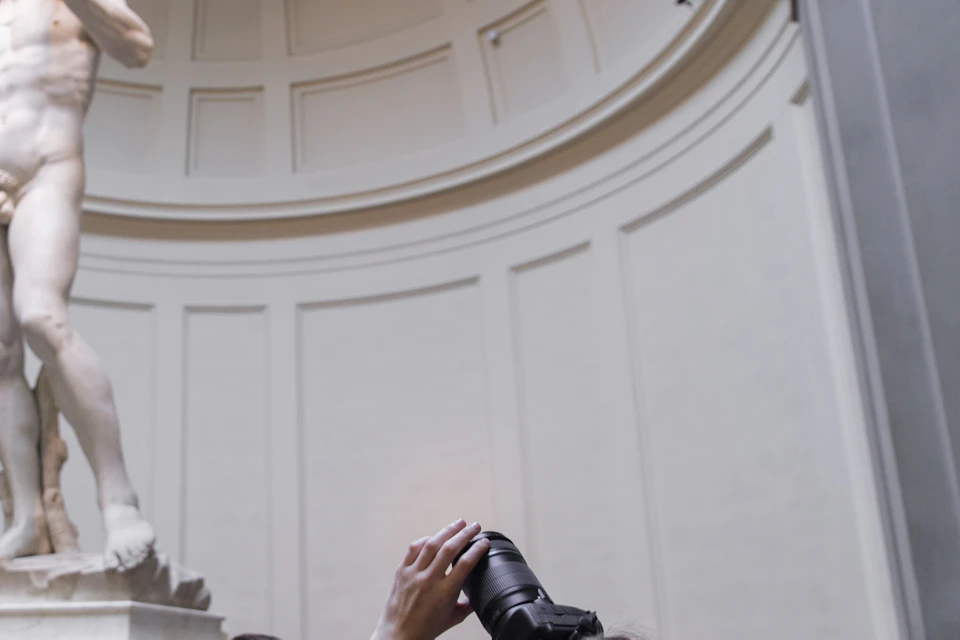A person photographs Michelangelo's David statue inside a museum while visitors observe the artwork.