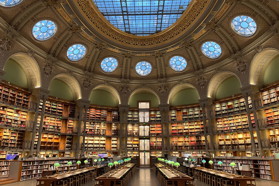 A spacious library with a domed glass ceiling, circular bookshelves and rows of tables with green reading lamps.