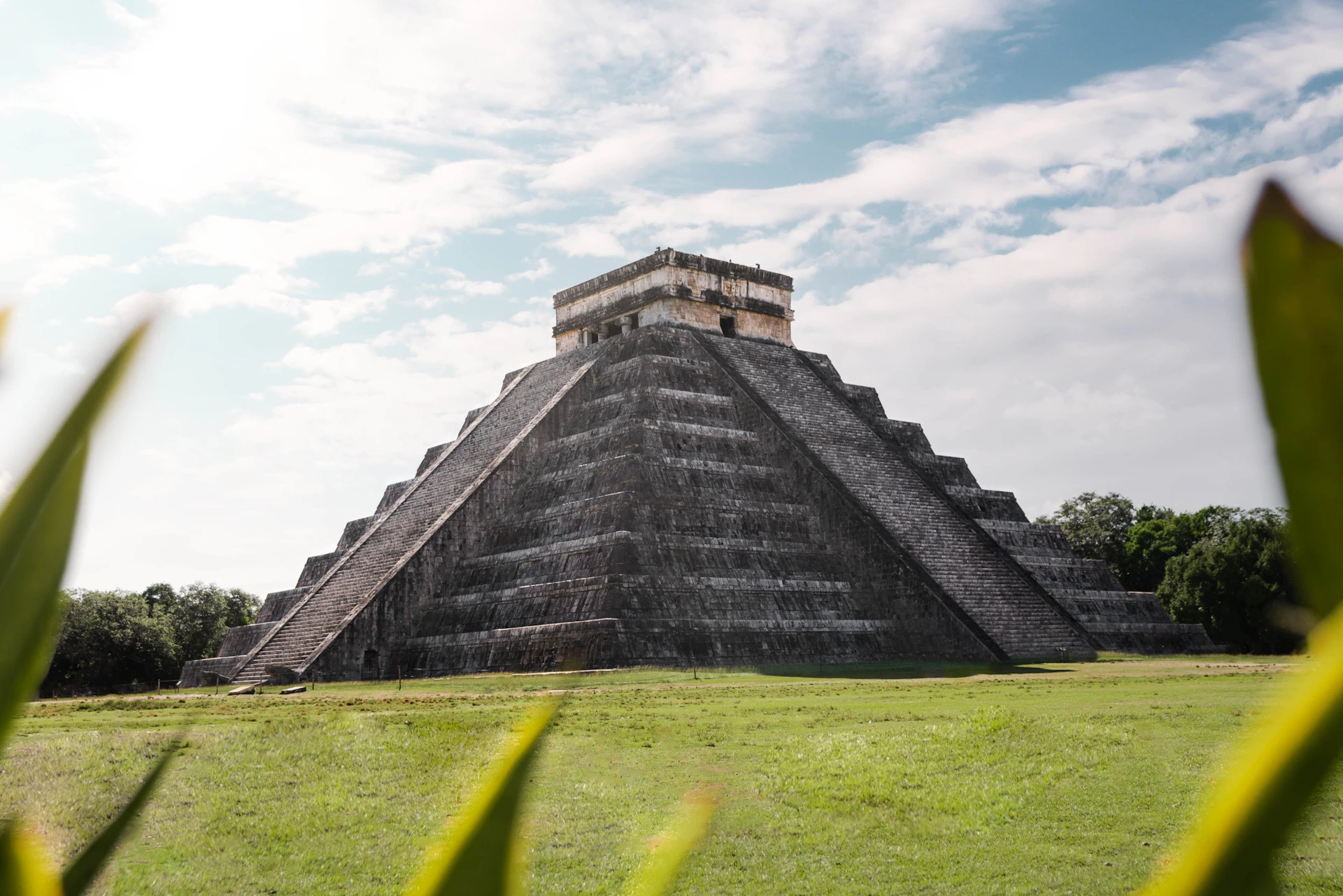 A large ancient stone pyramid with a staircase, set against a blue sky with scattered clouds, surrounded by green grass.