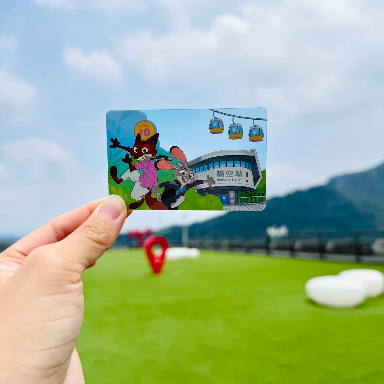 A hand holds a card depicting a cartoon scene with animals near a cable car station; a park and mountains are in the background.