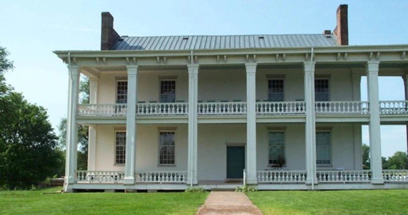 Two-story white building with large columns, balconies, and a metal roof. A dirt pathway leads to the entrance. Green lawn in front.