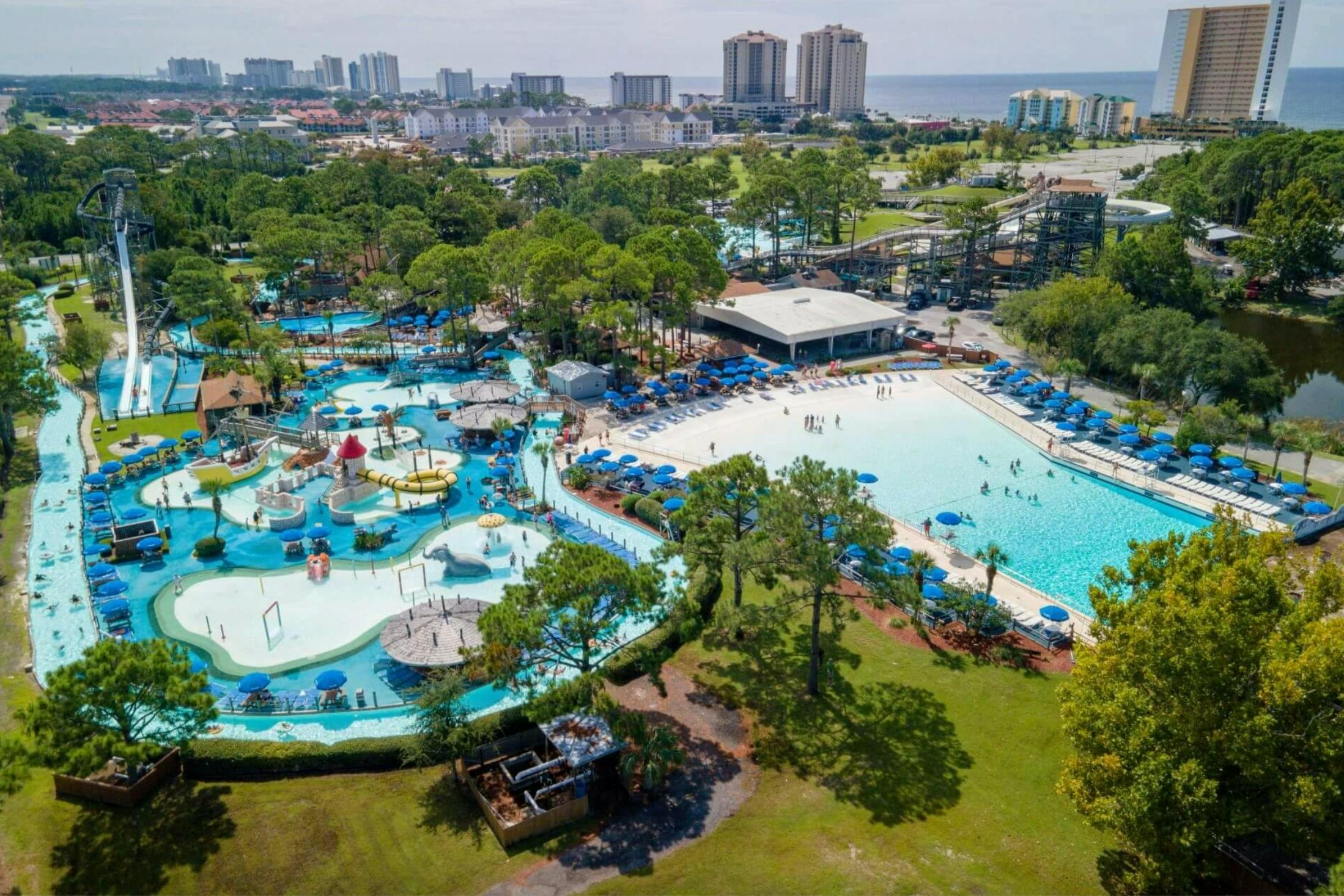 Vista aèria d'un parc aquàtic amb piscines, tobogans i para-sols envoltat d'arbres i edificis al fons.