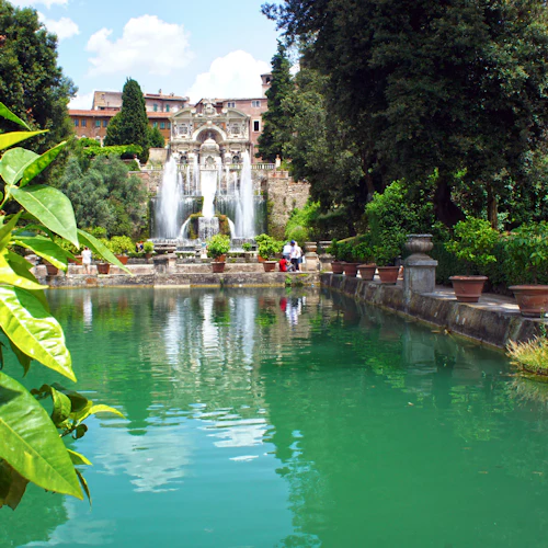 Grön damm med överhängande löv, krukväxter längs kanten och kaskaderande fontäner med statyer i en trädgårdsmiljö.