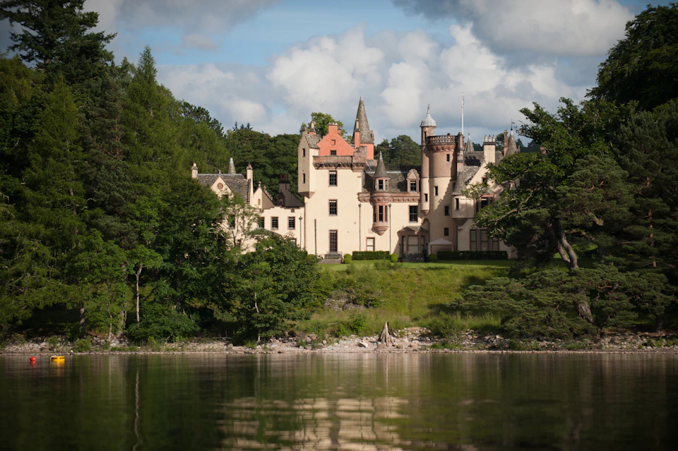 Cream-colored castle with turrets and steep roofs, surrounded by lush greenery, reflected in a calm body of water.