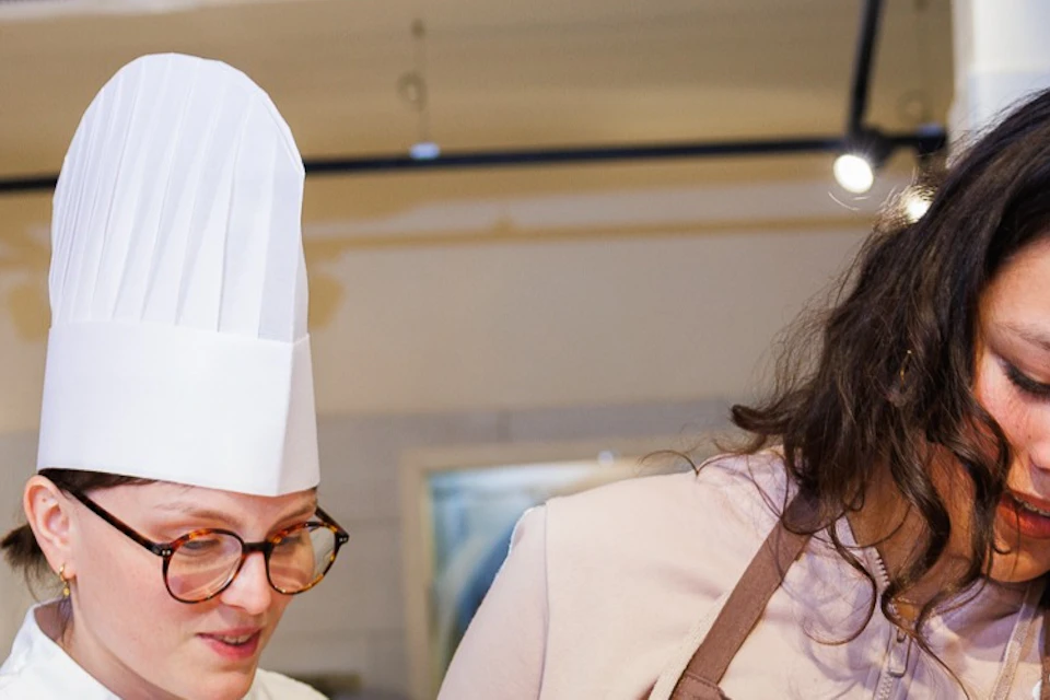 A group of people in aprons and a chef are decorating chocolates at a workshop. Bowls of ingredients are on the table.