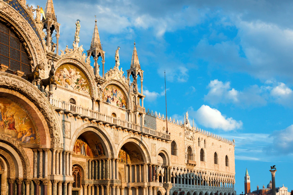 Ornate, historic building with arches, intricate carvings, statues, and mosaic artwork under a blue sky with scattered clouds.
