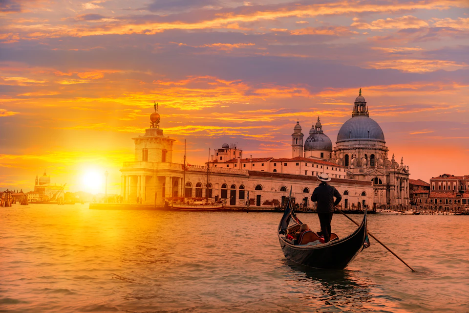 A person steers a gondola at sunset, passing historic domed buildings along a waterfront.