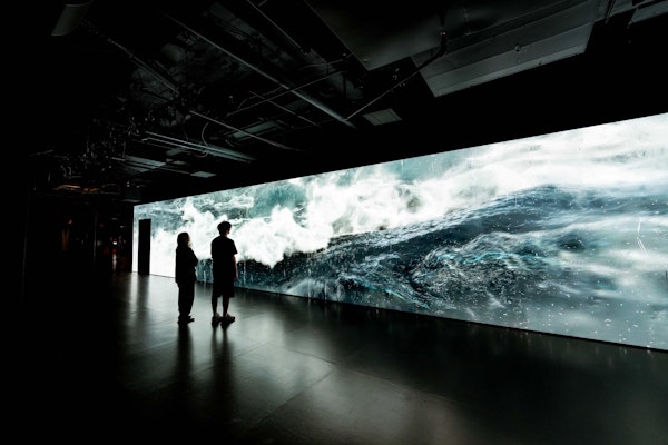 To personer står i et mørkt rum og ser på en stor digital skærm, der viser en turbulent havscene.