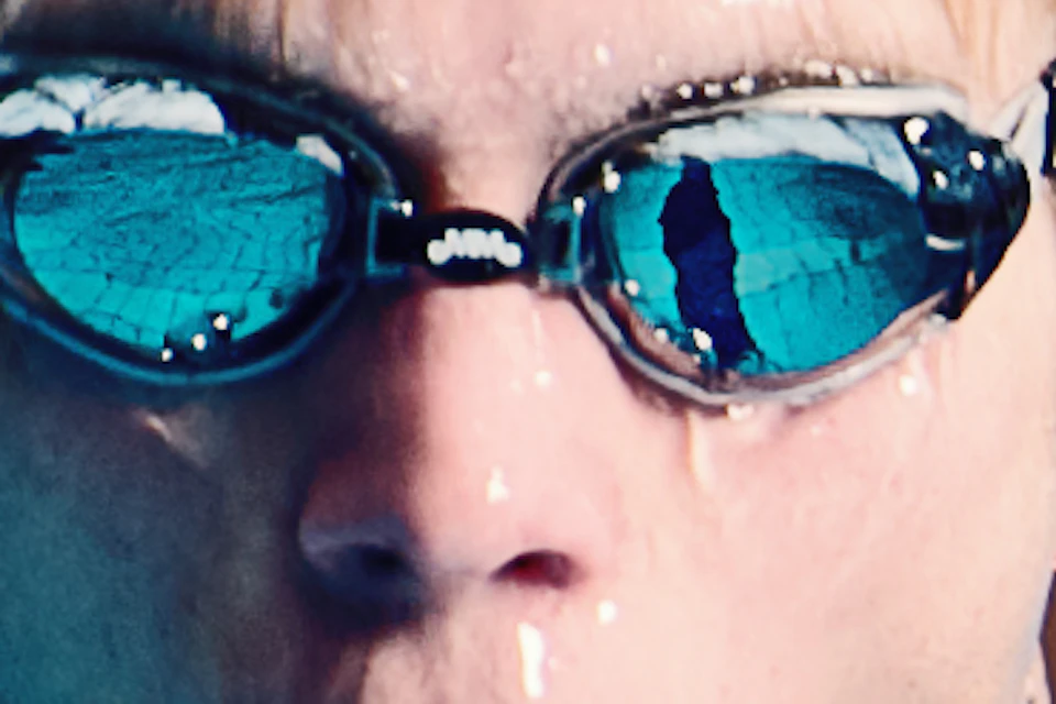 A swimmer with goggles emerges from a pool, water splashing around, with a person sitting in the background.