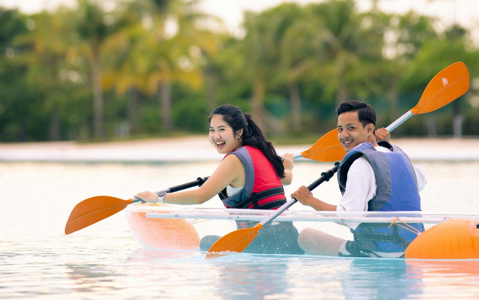 Two people paddling a transparent kayak on calm water, smiling, with trees and a beach visible in the background.