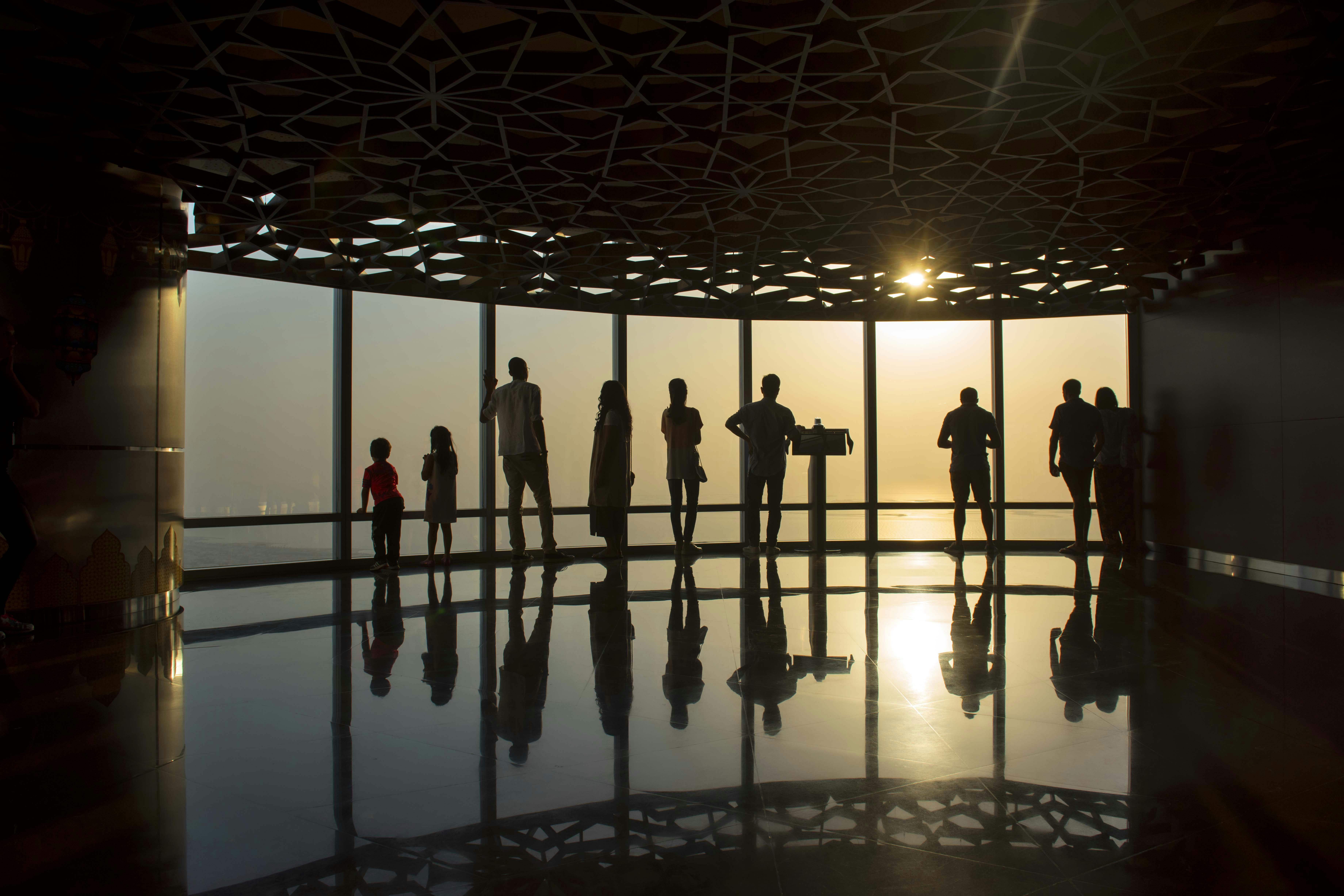 Silhouetten van mensen staan bij zonsondergang bij grote ramen, in een kamer met een plafond met patronen en een spiegelende vloer.