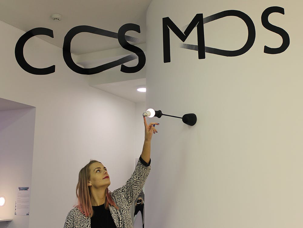 一个房间里，一个女人伸手去摸一个点燃的壁灯，墙上挂着一个大大的黑色字母，拼写着“COSMO”。