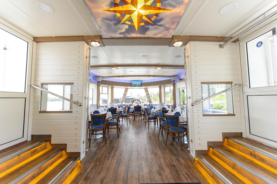 Elegant dining area on a boat with wooden floors, blue-cushioned chairs, and nautical decor, featuring a bright ceiling compass.