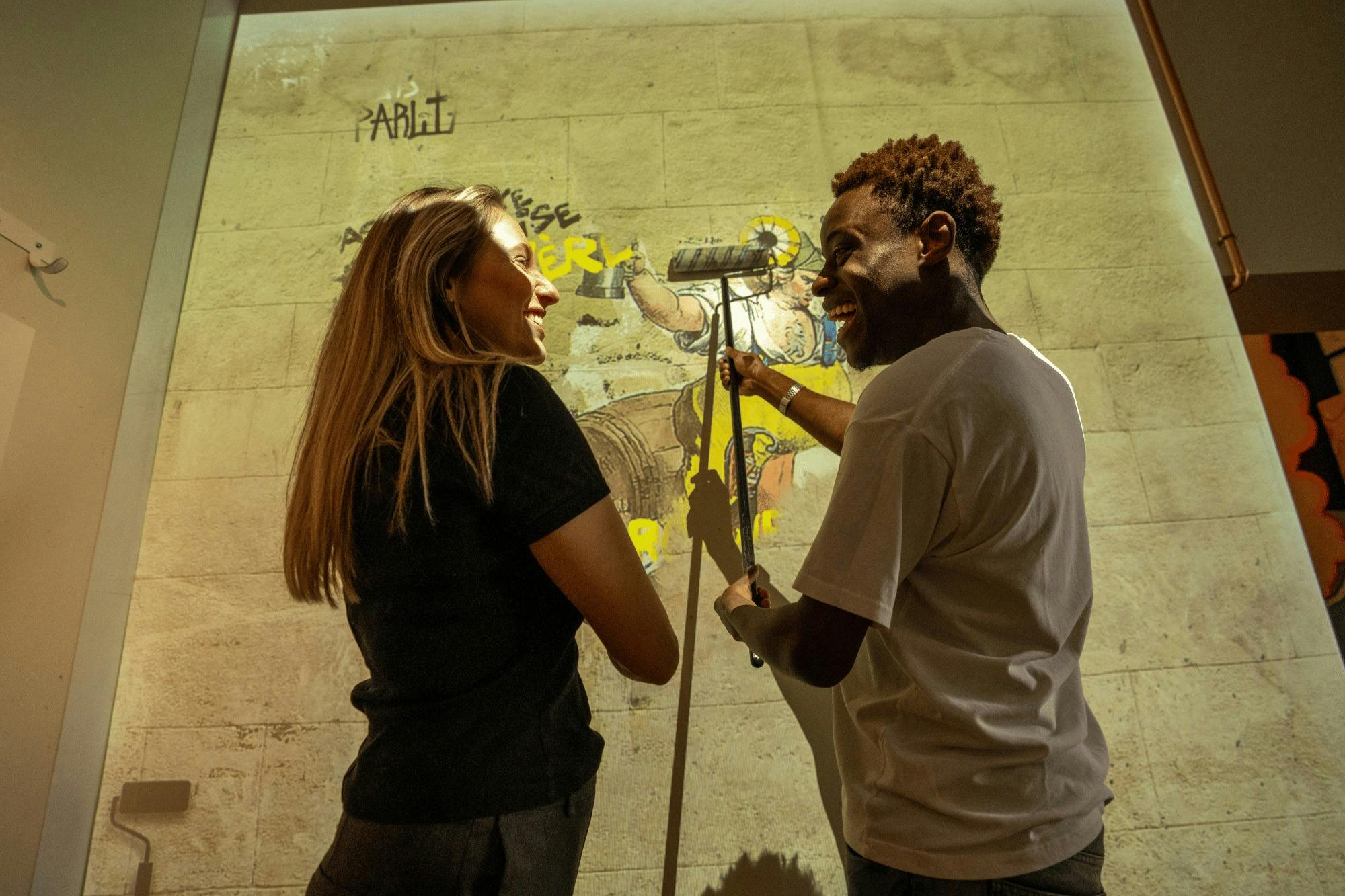 两个人，一个女人和一个男人，站在室内的插图墙前，微笑着投射。