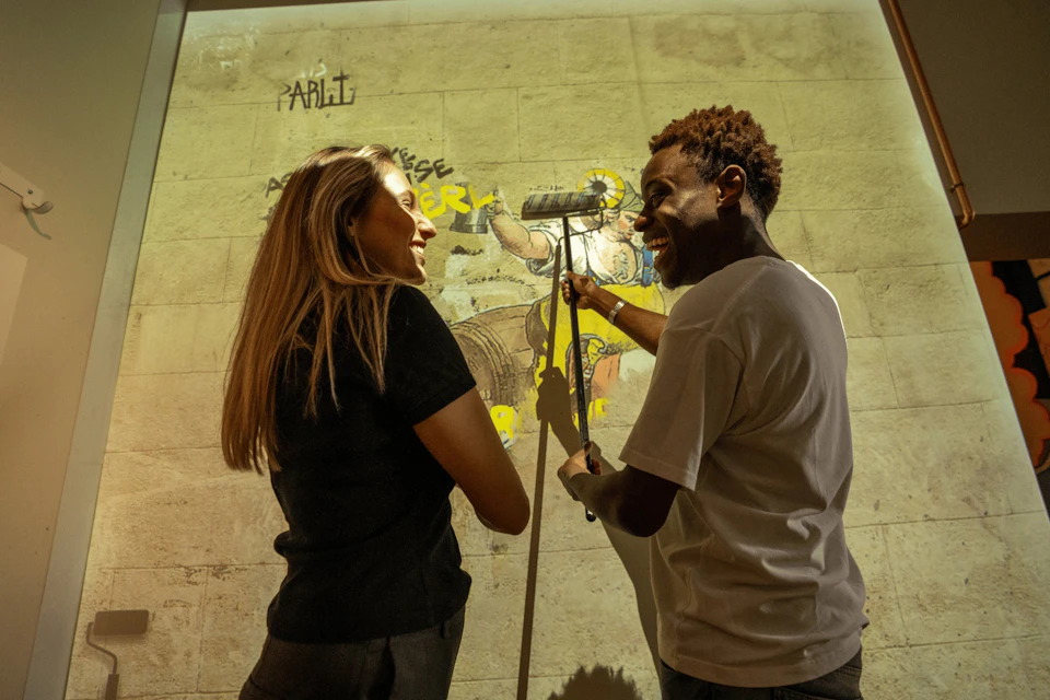 Two people, a woman and a man, stand indoors in front of an illustrated wall, smiling and engaging with a projection.