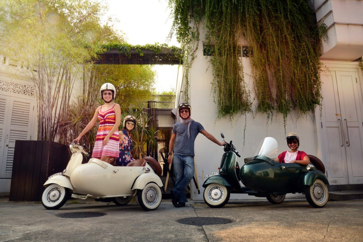 Trois personnes portant des casques se tiennent à côté de scooters anciens. L'un des scooters est équipé d'un side-car. L'arrière-plan est composé de plantes et d'un bâtiment blanc.