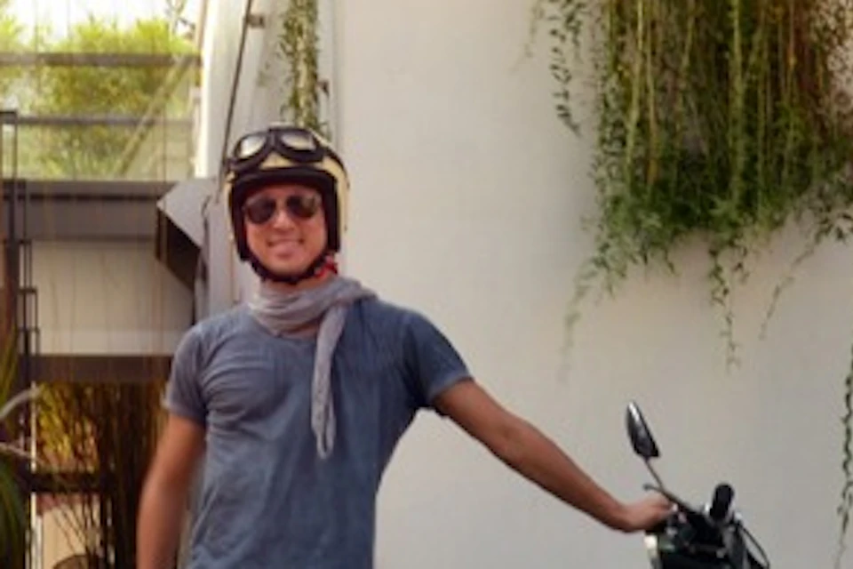 Three people with helmets are standing next to vintage scooters. One scooter has a sidecar. The background has plants and a white building.