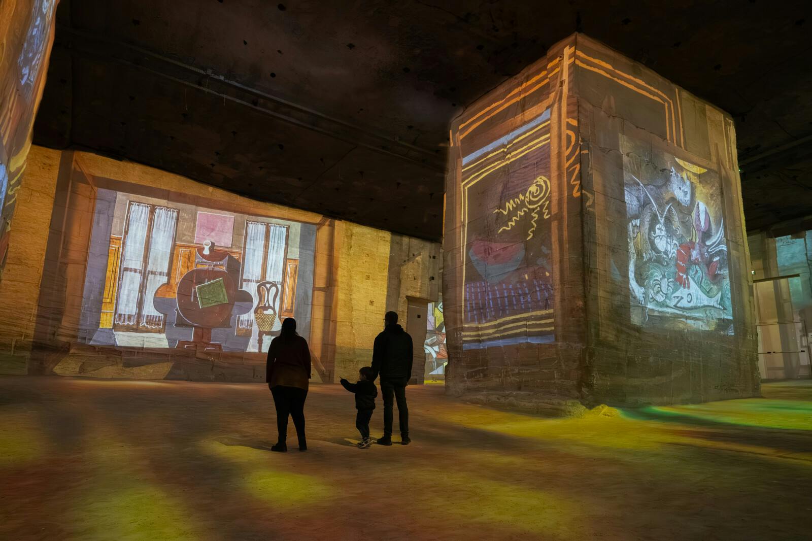 Three people observe illuminated artwork projections on large walls in a dimly lit gallery space.