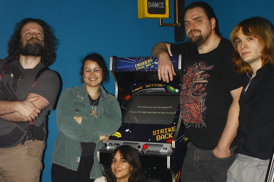 A group of people posing around a retro arcade machine with a space-themed design in a room with a blue wall.