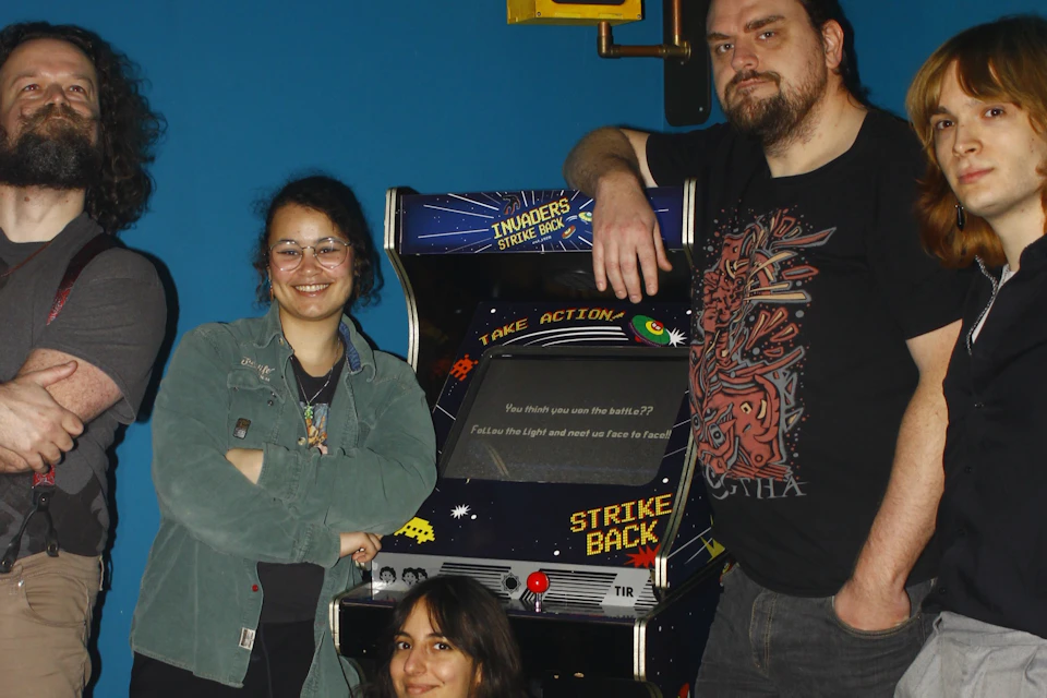 A group of people posing around a retro arcade machine with a space-themed design in a room with a blue wall.