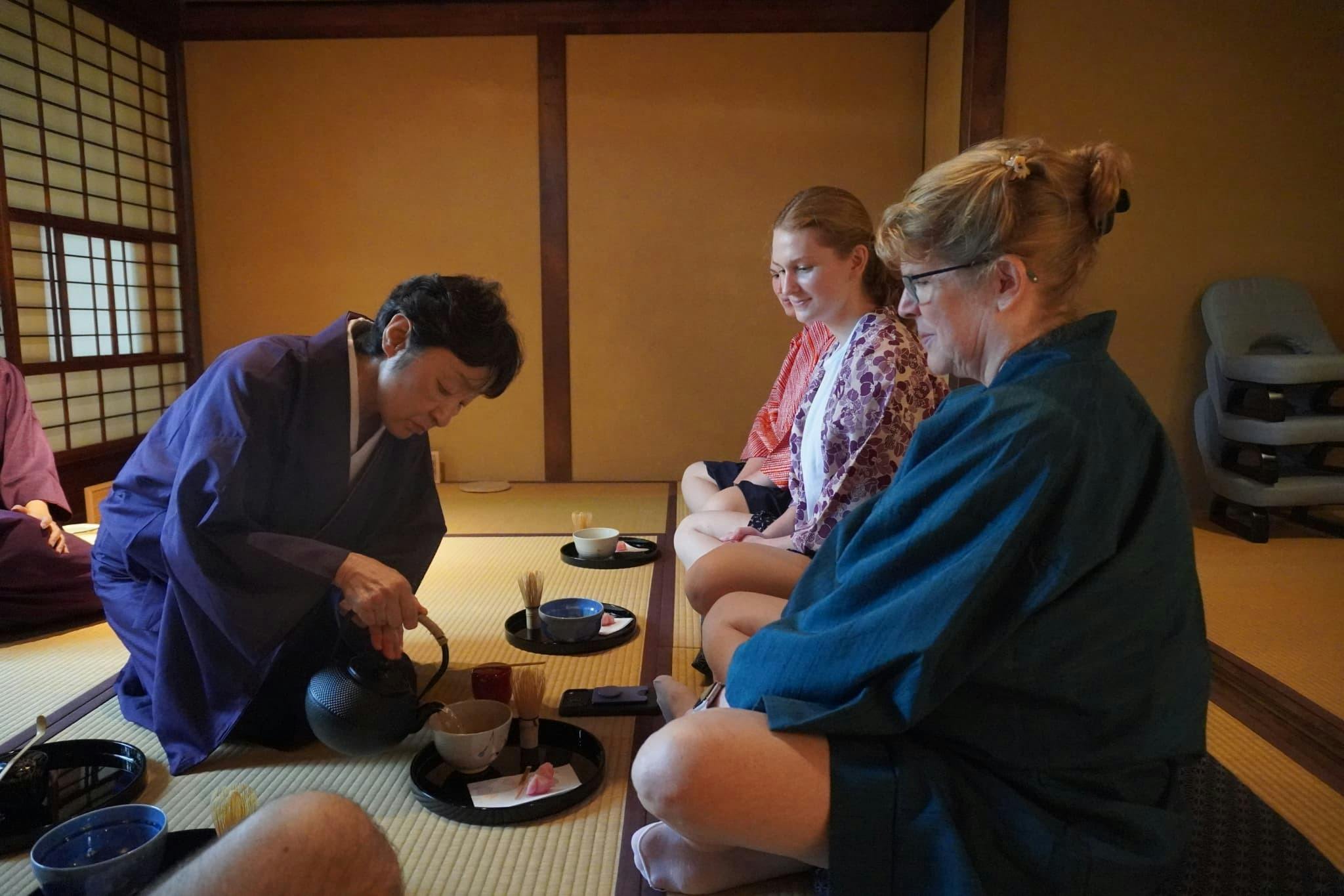 Three people seated on tatami mats, participating in a tea ceremony, with traditional Japanese bowls and utensils.