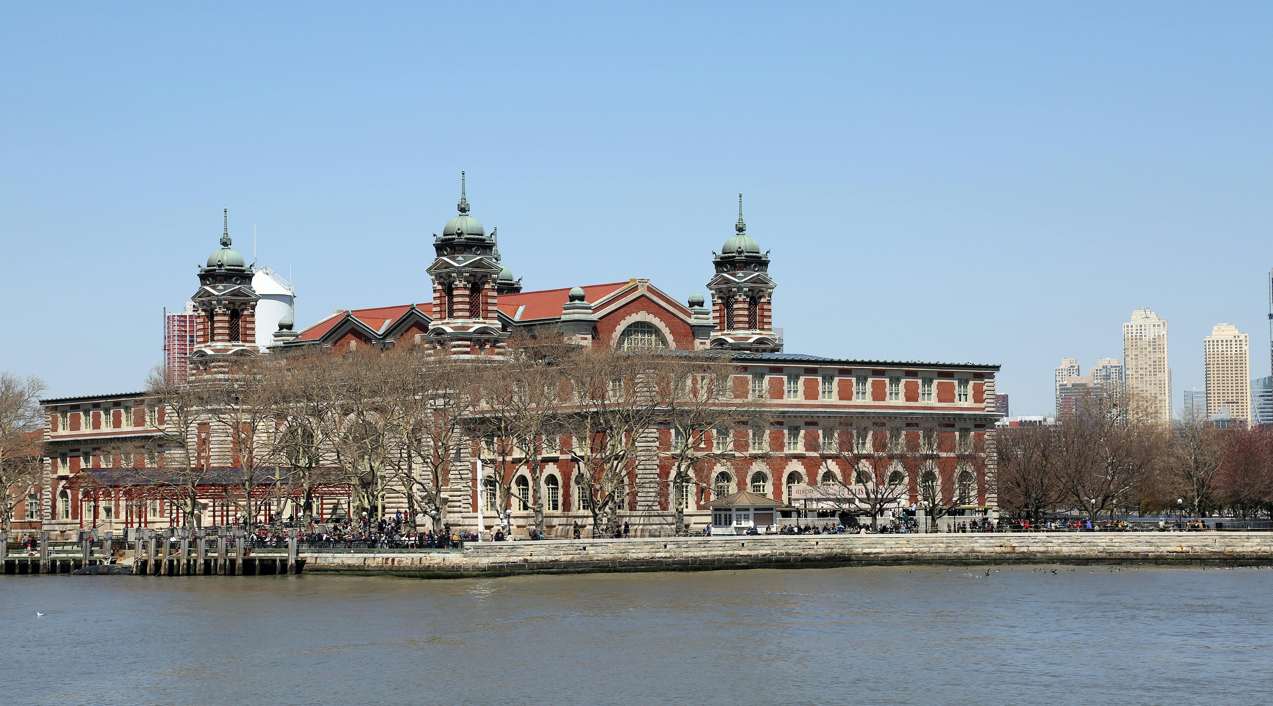 Un edificio in mattoni rossi con due torri gemelle e un lungomare, con alberi senza foglie e gente che si aggira in una giornata di sole.
