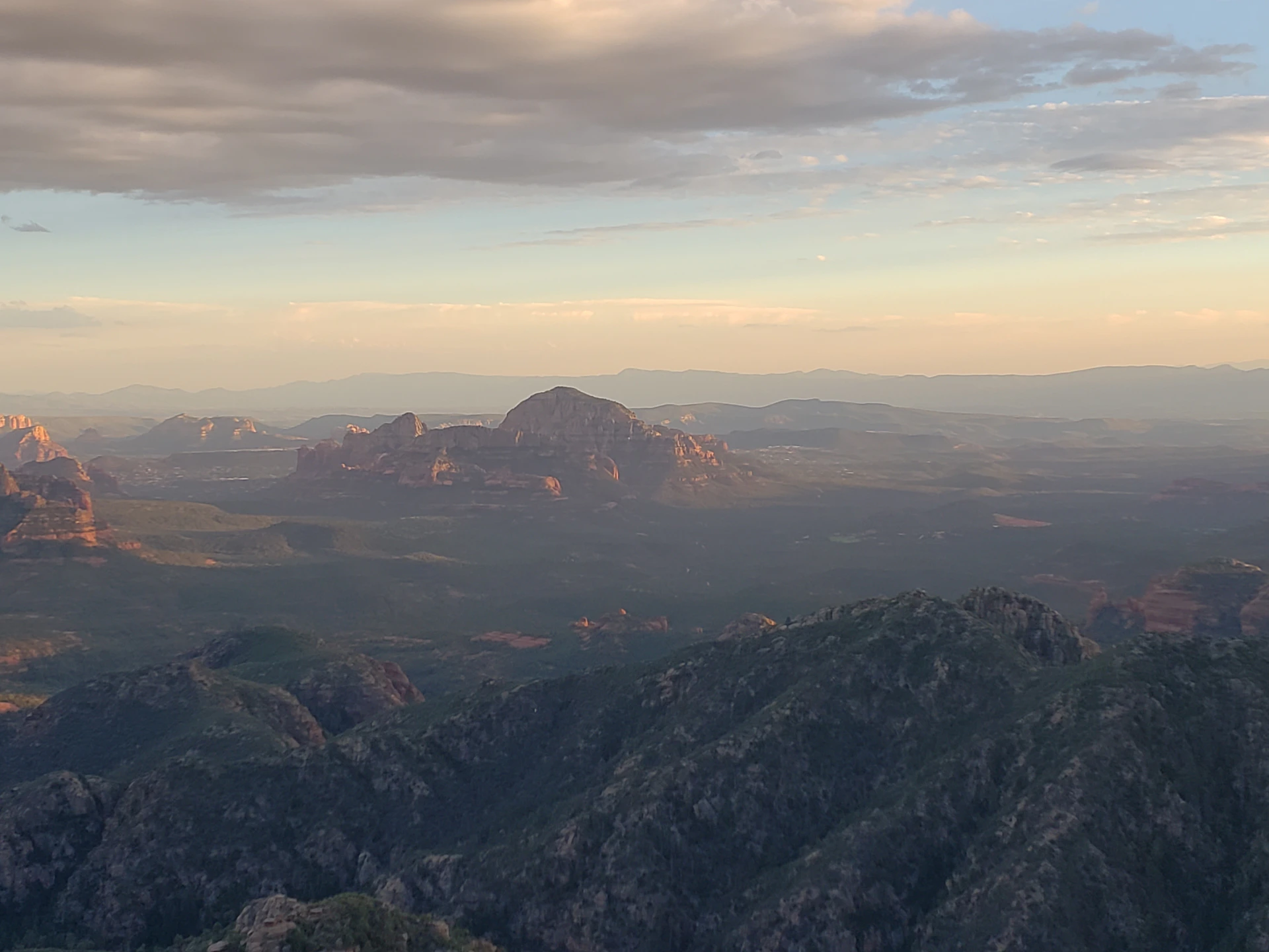 Mountainous landscape with rocky peaks, a distant mesa, and a sky filled with clouds during sunset.
