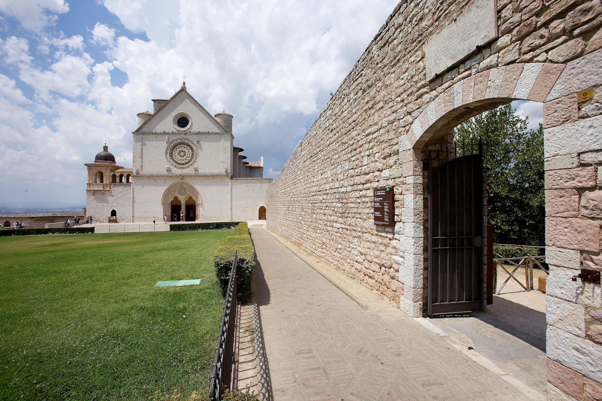 Stone pathway leading to a white church with a circular window, green lawn on the left, and arched gate entrance on the right.