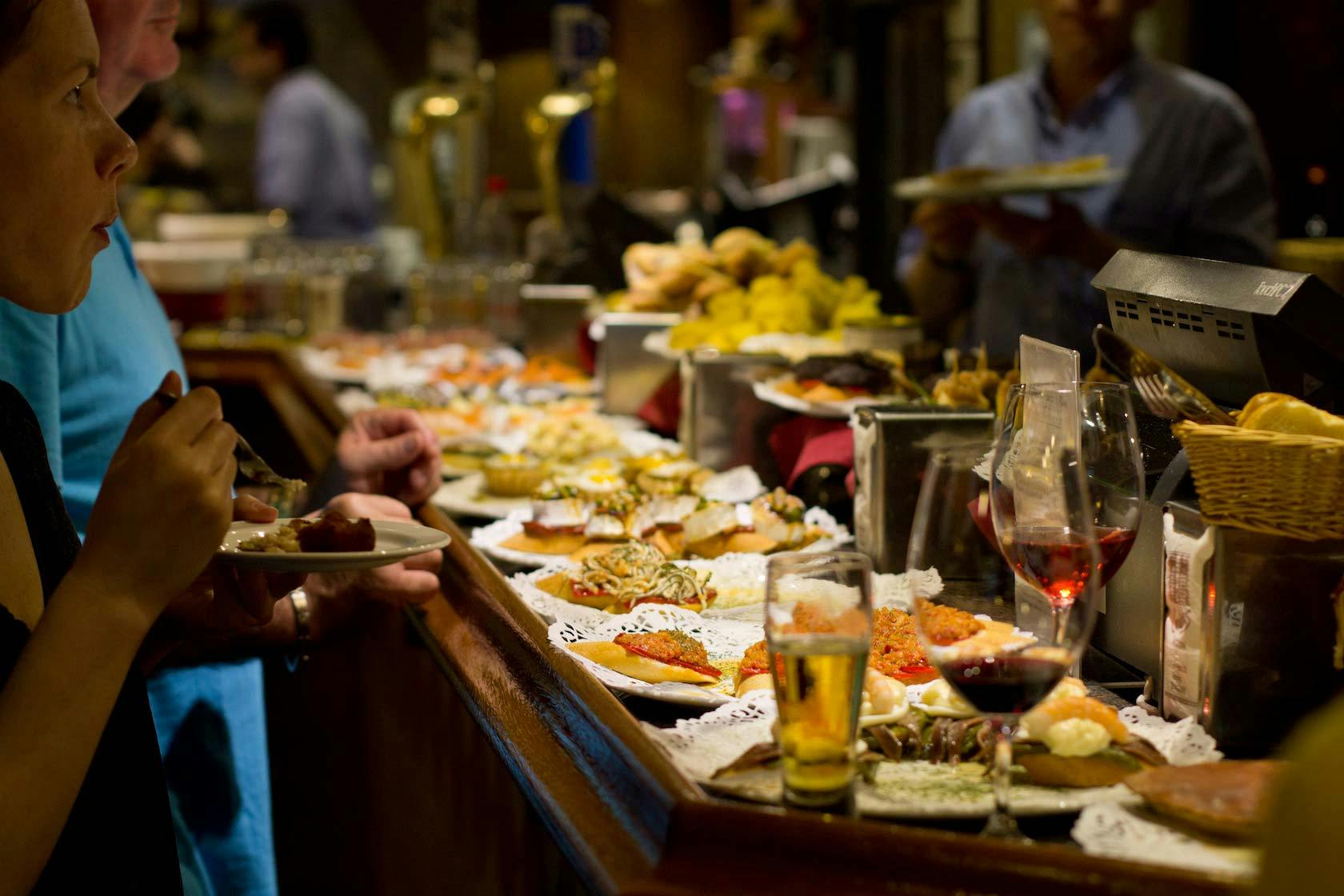 酒吧或塔帕斯餐厅的人们正在享用各种小菜小饮，柜台上摆放着餐盘和玻璃杯。