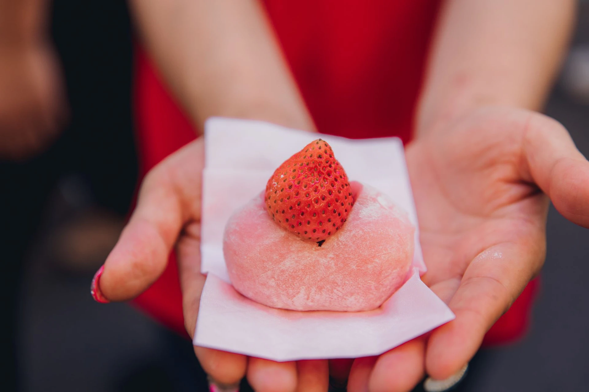 A person holding a pink mochi topped with a fresh strawberry on a piece of white paper.