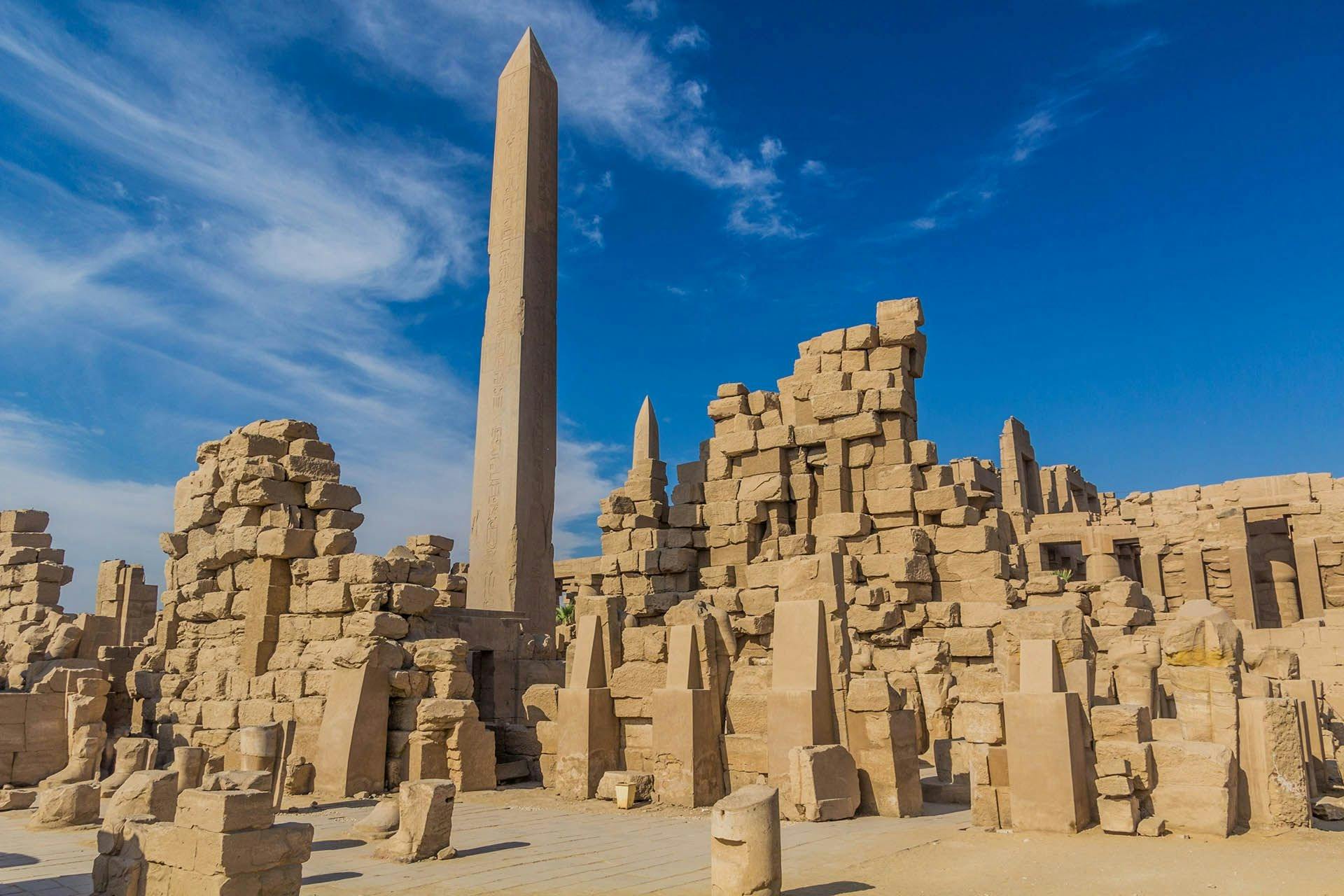 Ancient ruins with tall obelisks and scattered stone structures under a blue sky with light clouds.